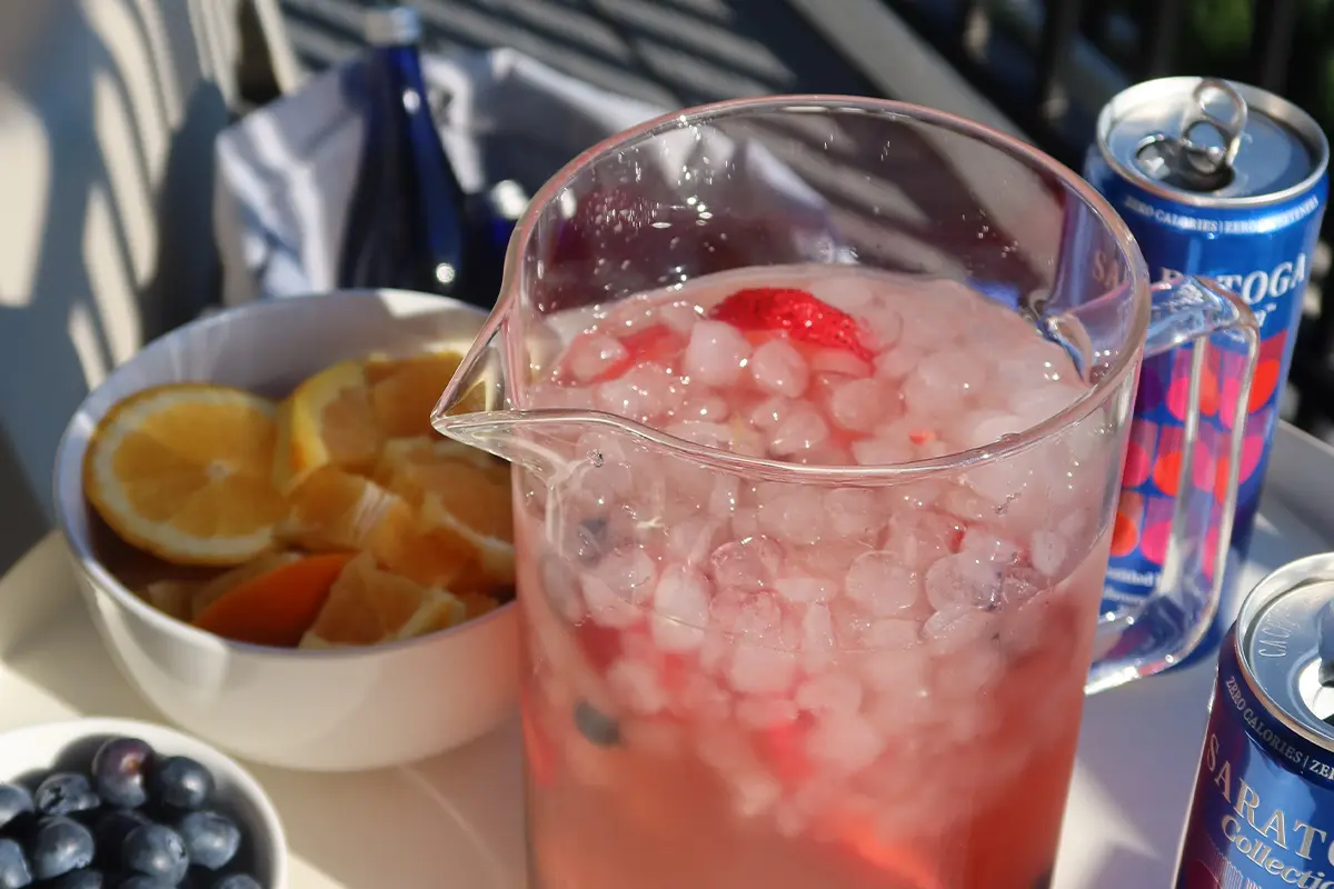 A pitcher of berry Sangria with berries and a strawberry, next to sliced oranges, blueberries, and soda cans on a sunlit table.