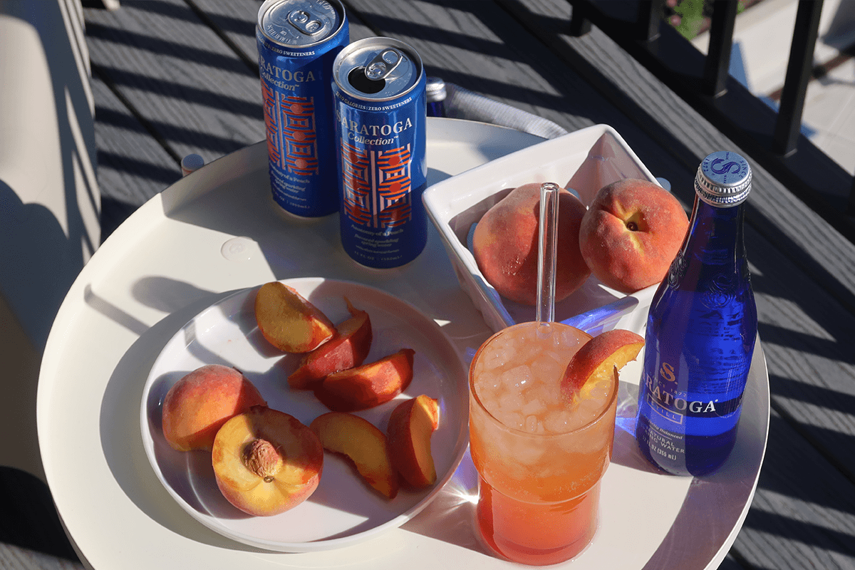 Peach slices on a plate, peaches in a bowl, a peach drink with ice, two cans, and a blue Saratoga bottle on a white table under sunlight.