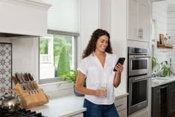 A woman in a kitchen, smiling while looking at her smartphone, holding a drink. Bright, modern kitchen with stainless steel appliances.