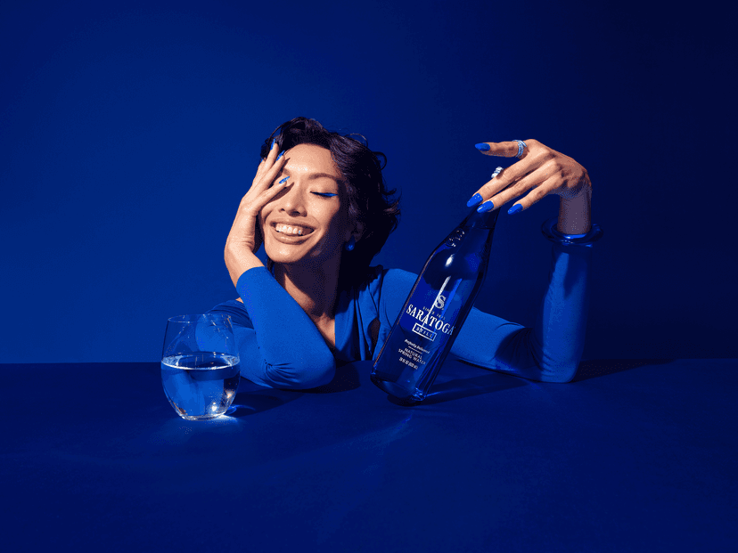 A person in a blue outfit leans on a table with a bottle of Saratoga sparkling water and a glass, smiling and resting a hand on their cheek.