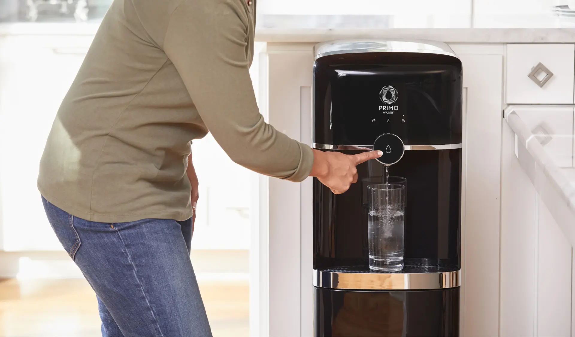A person pressing a button on a black Primo Water® water dispenser, filling a glass with purified water.