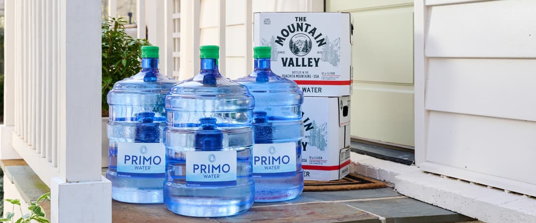 Large Primo water bottles and Mountain Valley boxes on a porch next to a white house wall and steps.