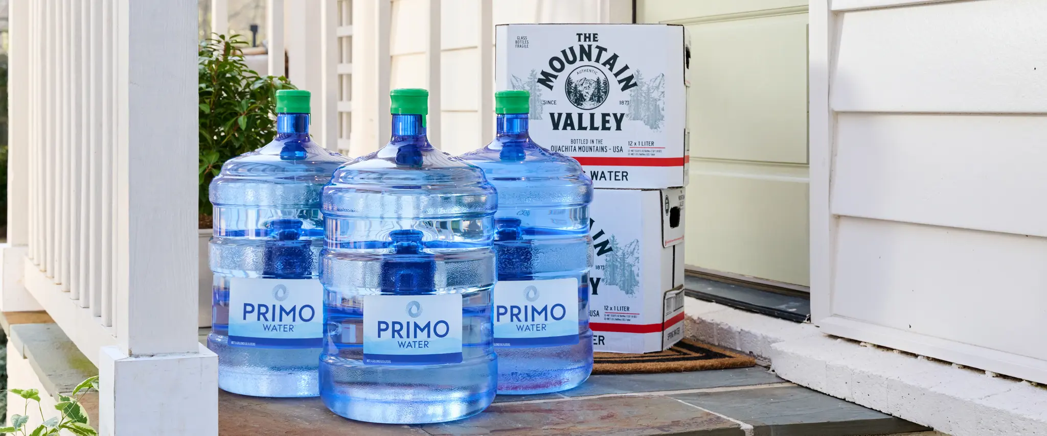 Large Primo water bottles and Mountain Valley boxes on a porch next to a white house wall and steps.
