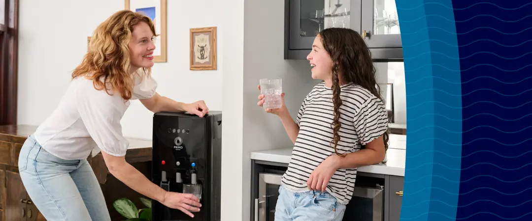 A woman fills a glass of water from a dispenser while a girl in a striped shirt and jeans leans against the counter, smiling with her own glass.