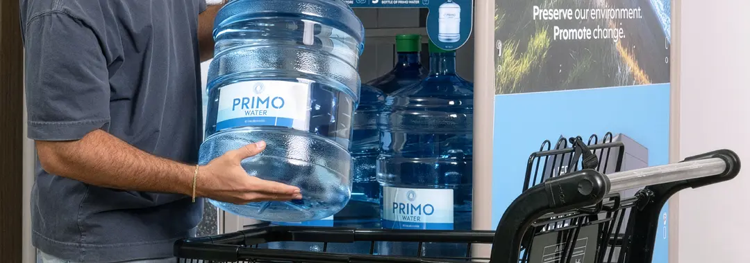 Person placing a large Primo water bottle into a shopping cart, with a backdrop of a Primo's exchange. station.