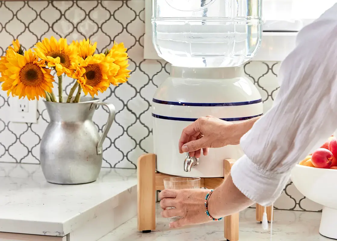 Counter top water dismenser next to a bowl of fruit and a pitcher of flowers