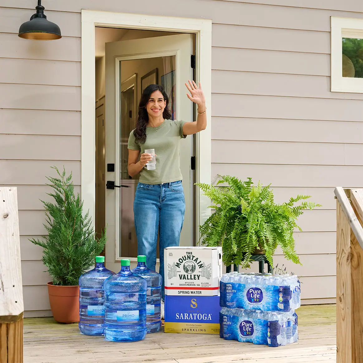 A woman in a green shirt and jeans stands at the doorway, waving with a drink in hand, surrounded by large water containers and potted plants.