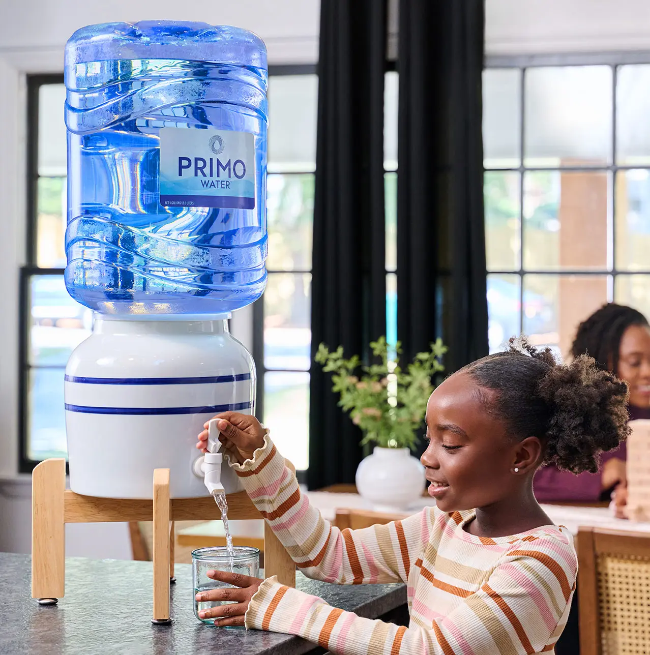 A young girl is filling a glass with water from a large blue Primo water dispenser. She is smiling, and a woman is blurred in the background.