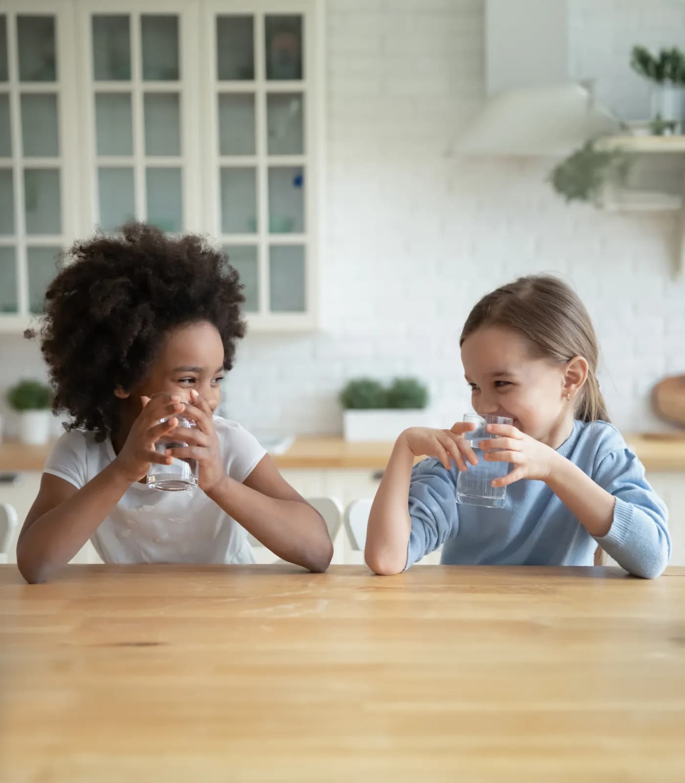 Two young girls smiling and drinking water at a kitchen counter