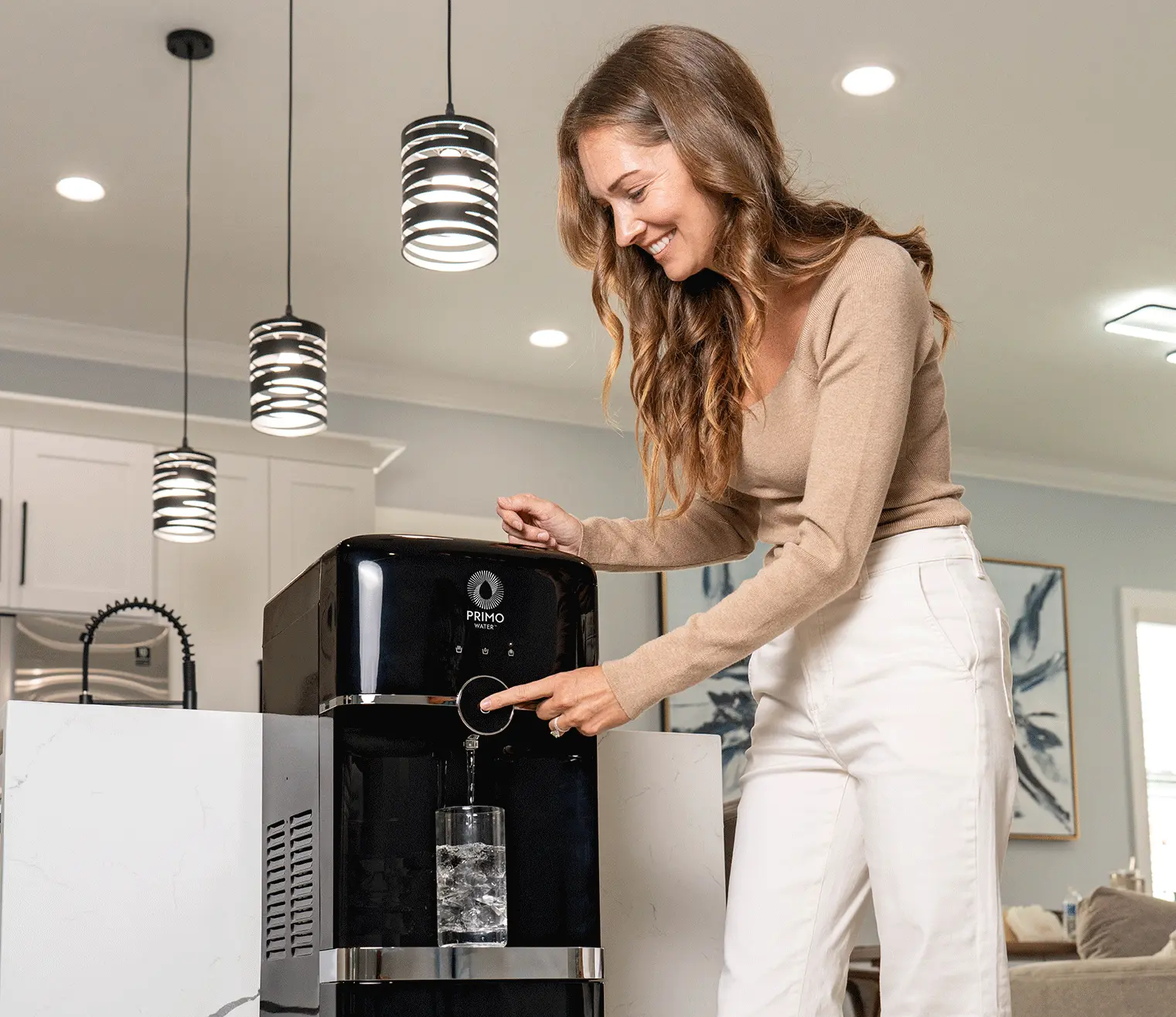 A woman in a kitchen dispenses water into a glass with ice from a Black Primo countertop water dispenser, smiling under modern pendant lights.