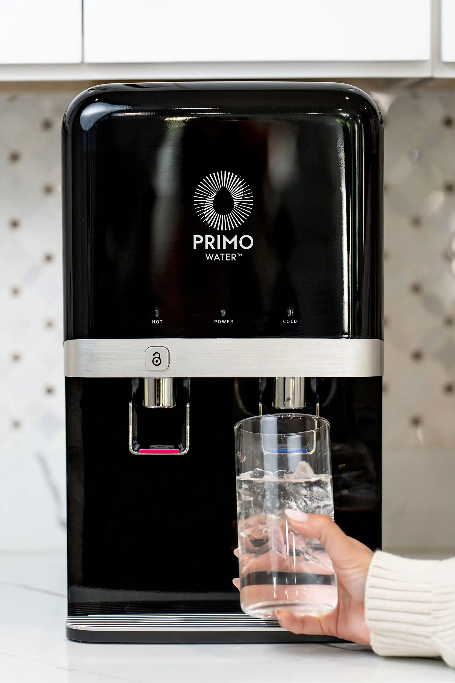 A person filling a glass with water from a black and silver Primo branded dispenser on a kitchen countertop.
