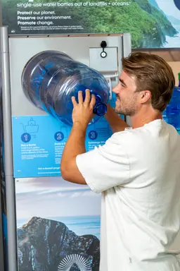 A man refills a large blue water jug at a water refill station with signage promoting environmental protection by reducing single-use bottles.