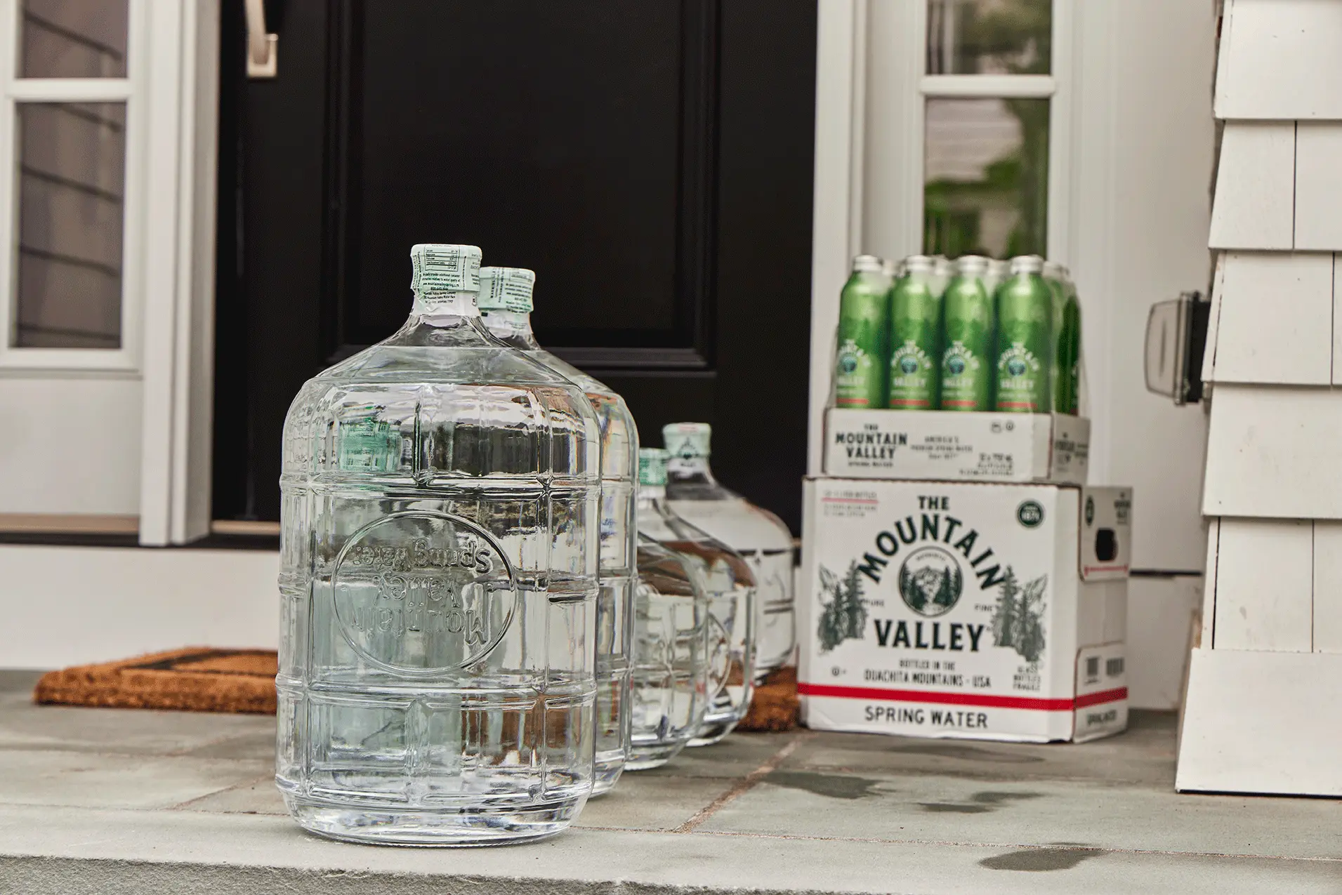Large water jugs and boxed cans of Mountain Valley spring water placed outside a front door on a stone step.