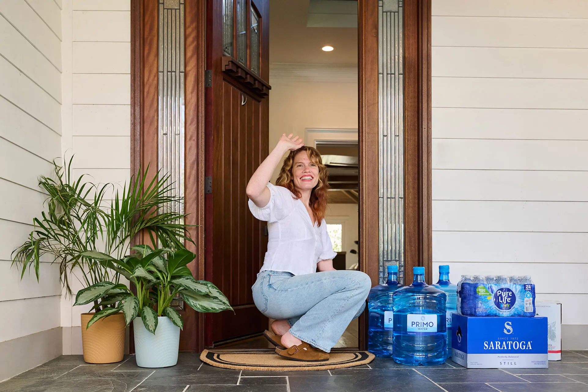 A woman kneels at her front door, smiling and waving, surrounded by potted plants and water bottles.
