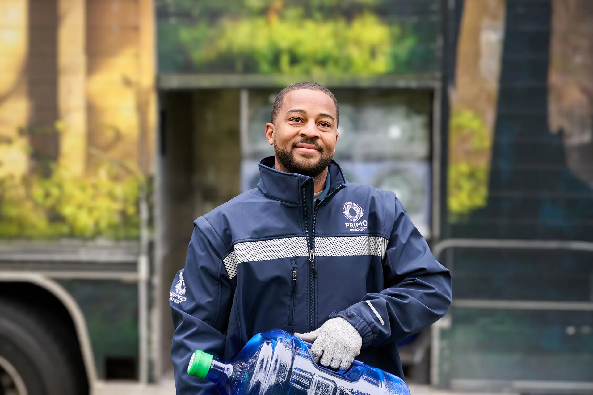 A person in a blue jacket with "Primo" logo, holding a large blue water bottle, stands outdoors with a mural in the background.