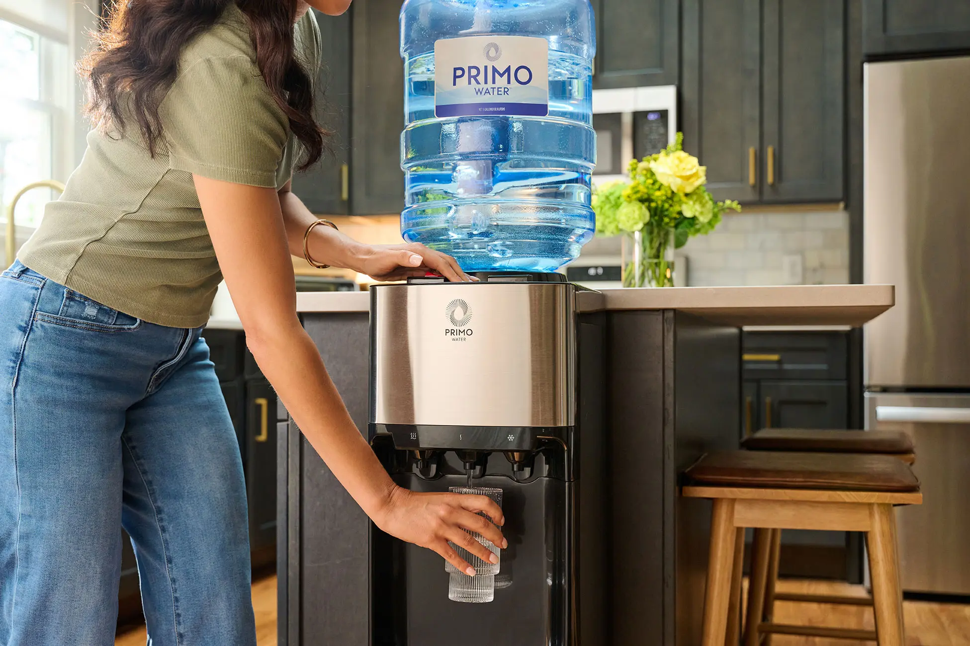A person filling a glass with water from a Primo water dispenser in a modern kitchen.