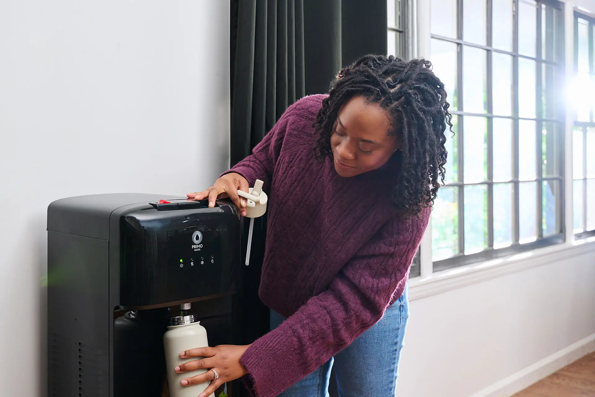 A person fills a bottle from a water dispenser while holding a white cap. Bright windows are in the background.