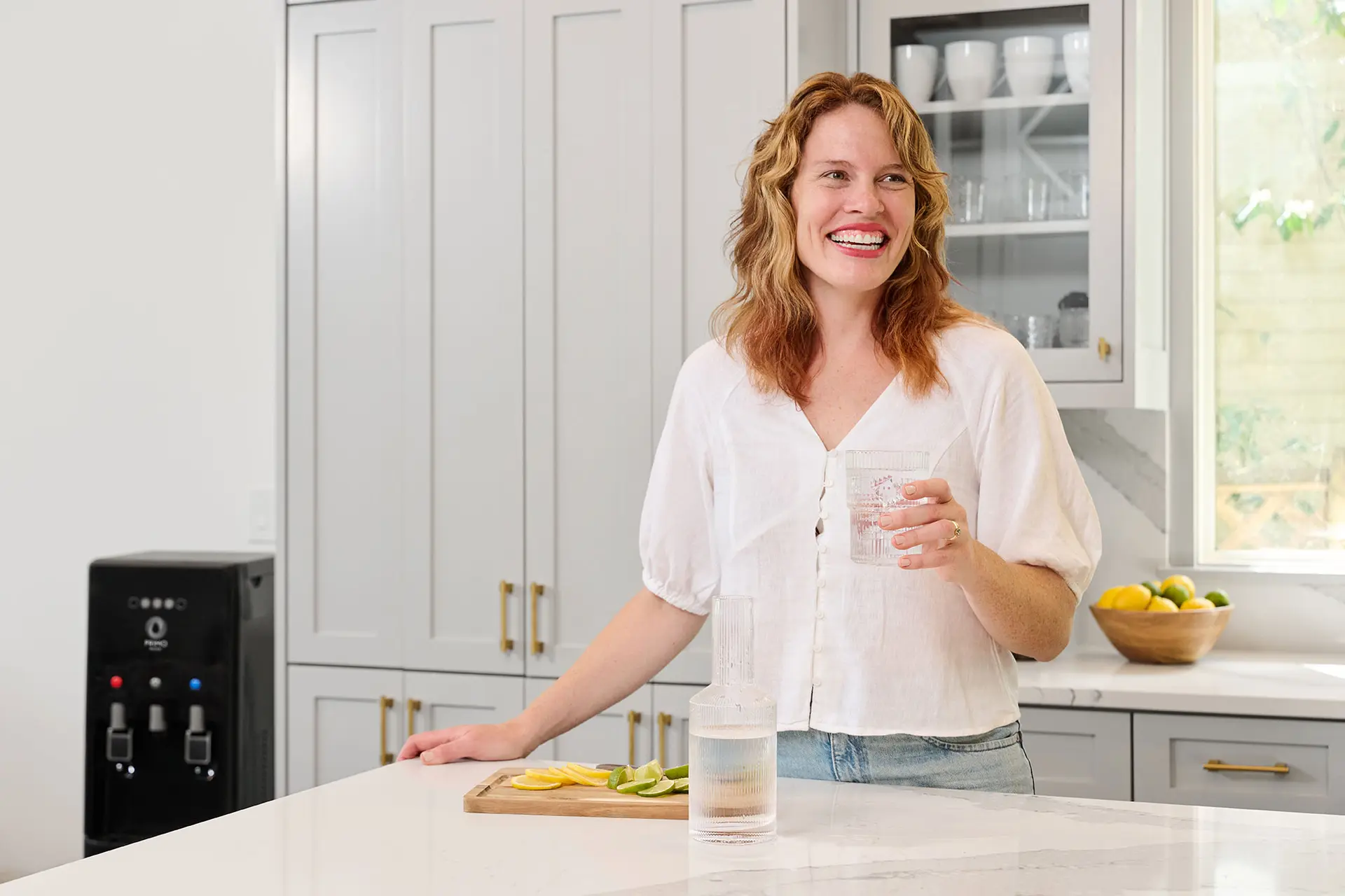 A woman in a white blouse stands in a modern kitchen, holding a glass of water beside a cutting board with sliced lemons and limes.