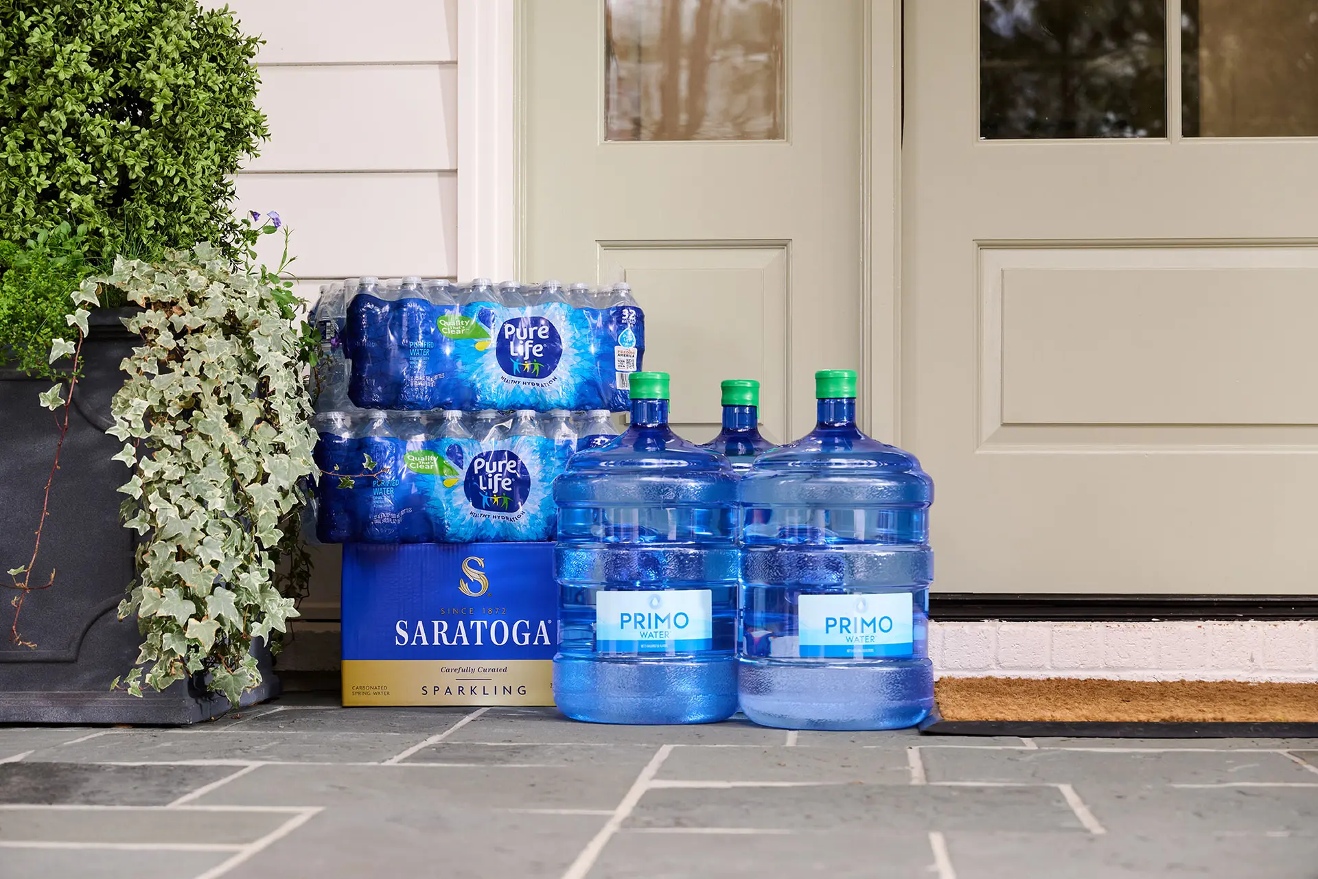 Cases of bottled water and large water jugs are stacked by a home's front door, next to a potted plant.