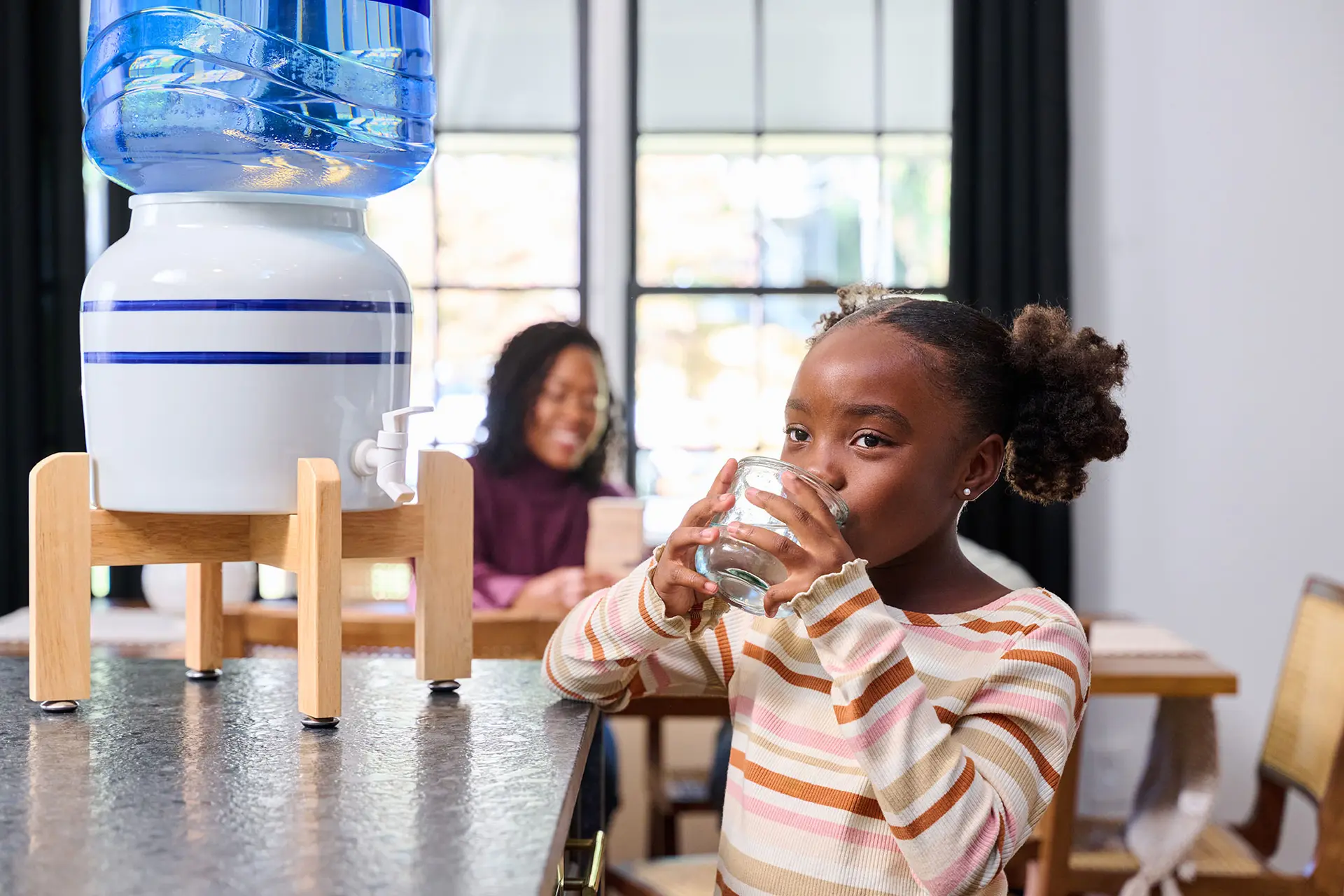 A young girl drinks water from a glass by a blue and white water dispenser while a woman sits in the background.