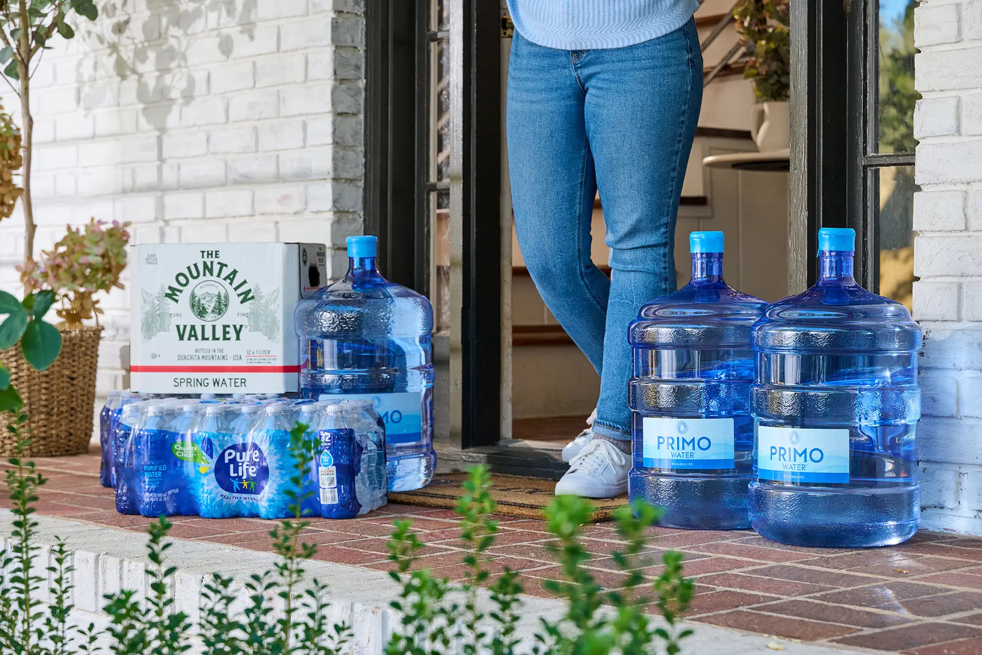 Person in jeans standing on a porch with large water jugs and bottled water packs nearby. Brick and potted plants visible.