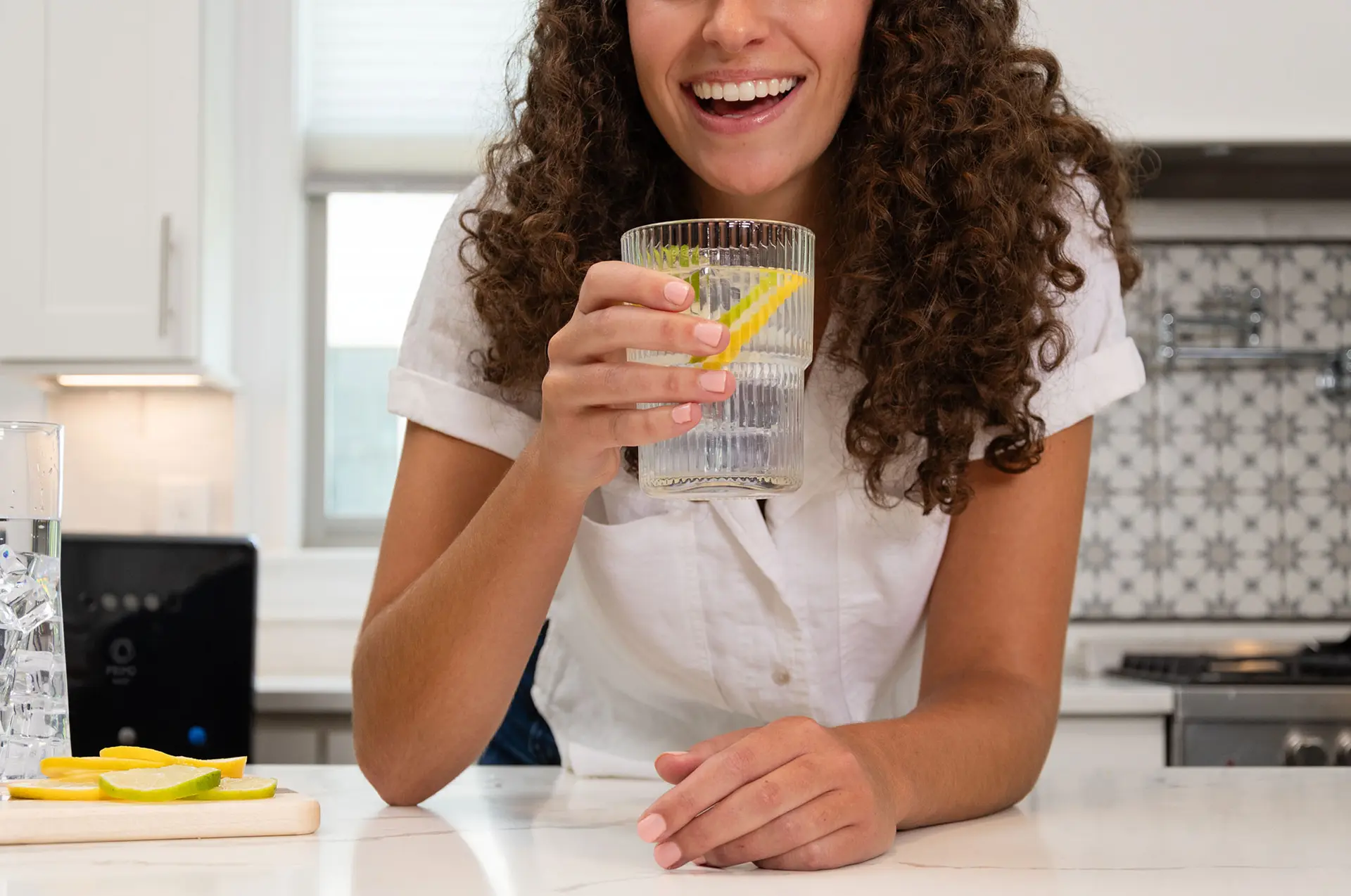 A smiling woman in a white shirt holding a glass with lemon slices in a bright kitchen.
