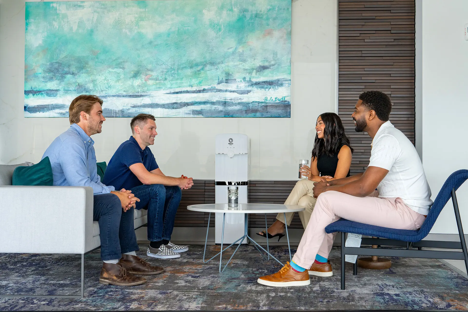 Four people sitting in an office lounge, engaged in conversation, with a colorful abstract painting in the background.