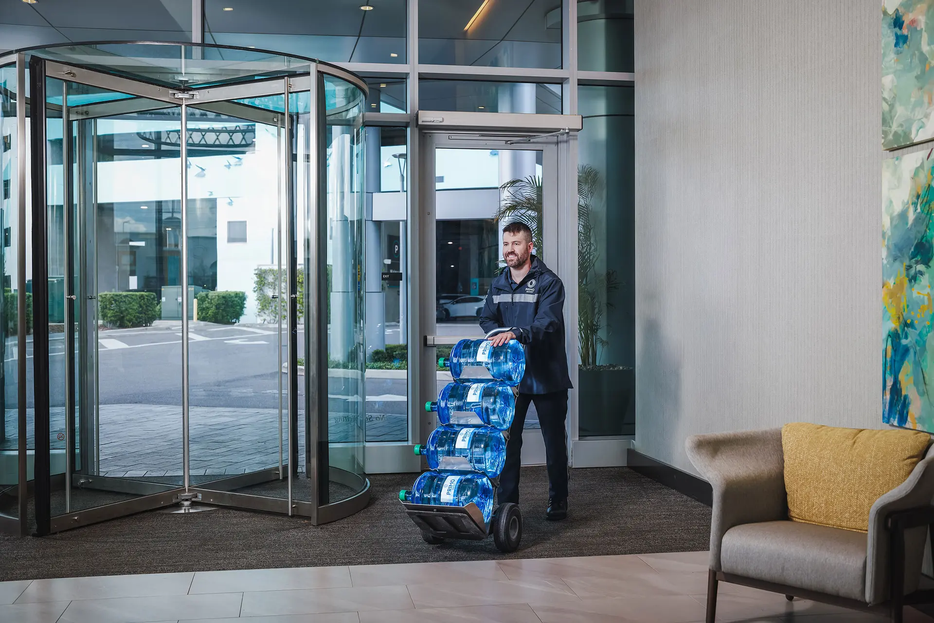 Man pushing a cart with four large water bottles into a modern building with a glass revolving door.