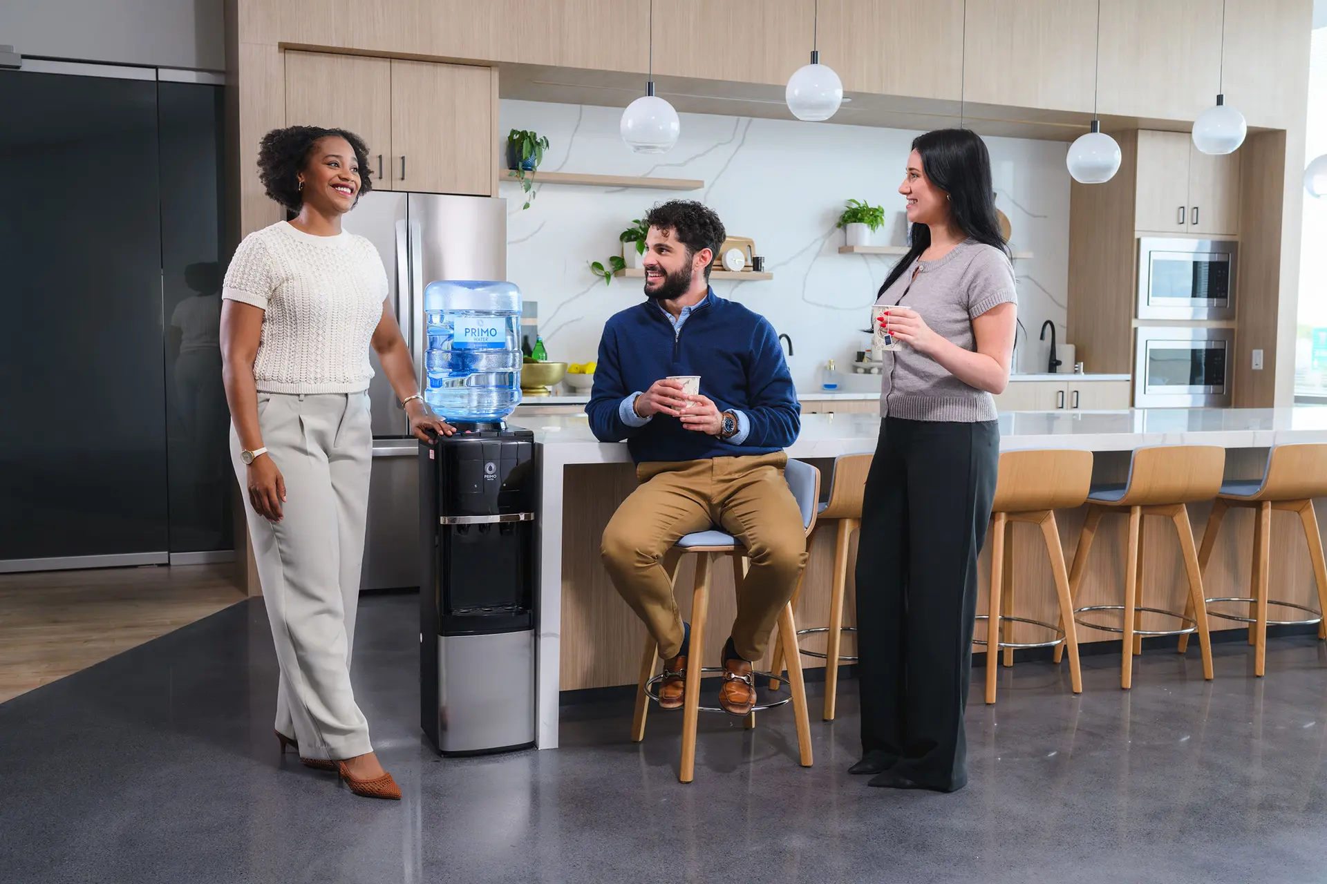 Three people talking and drinking coffee around a water cooler in a modern office kitchen with wooden cabinets and bar stools.