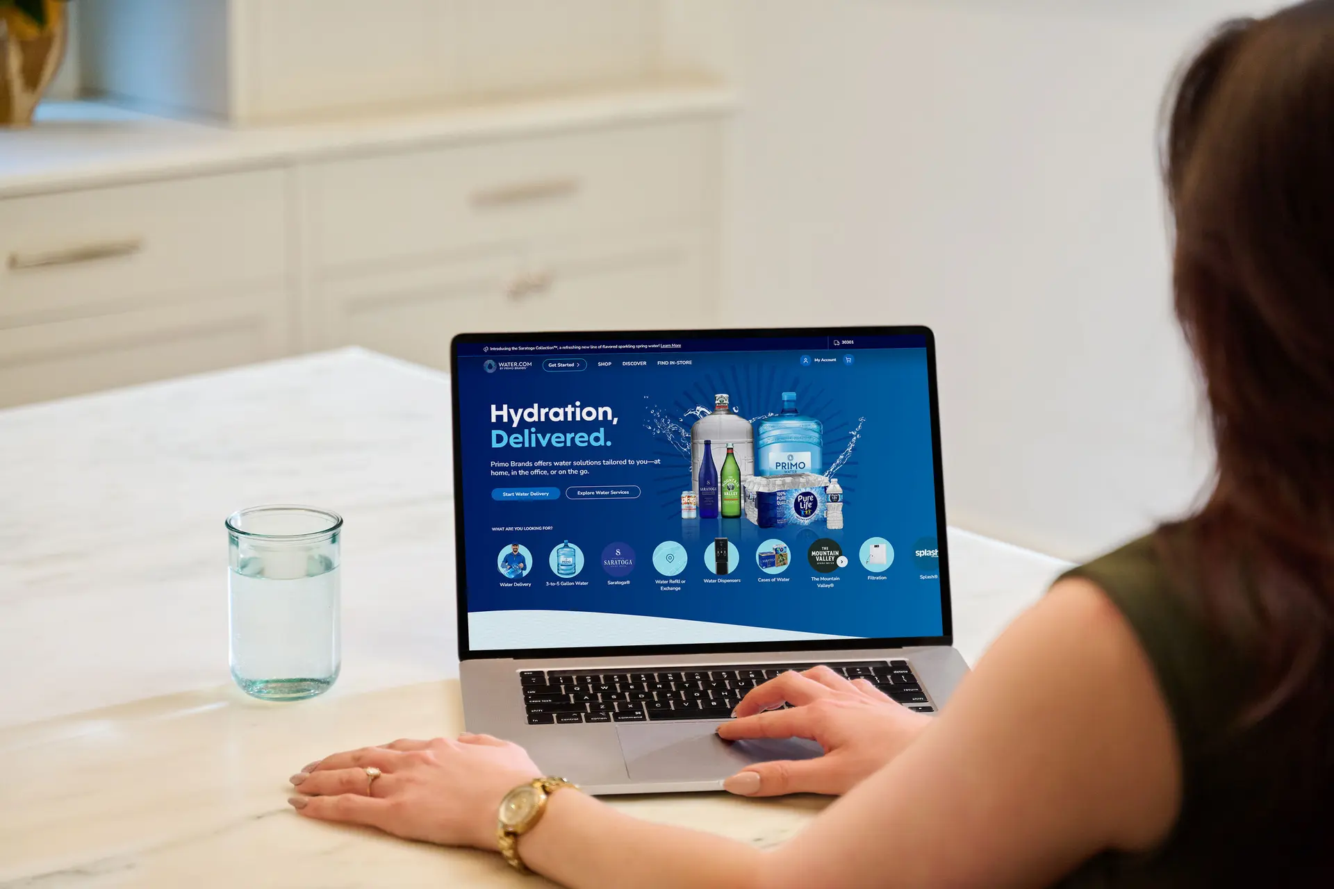 A person looks at a laptop displaying a water delivery service website. A glass of water is nearby on a white countertop.