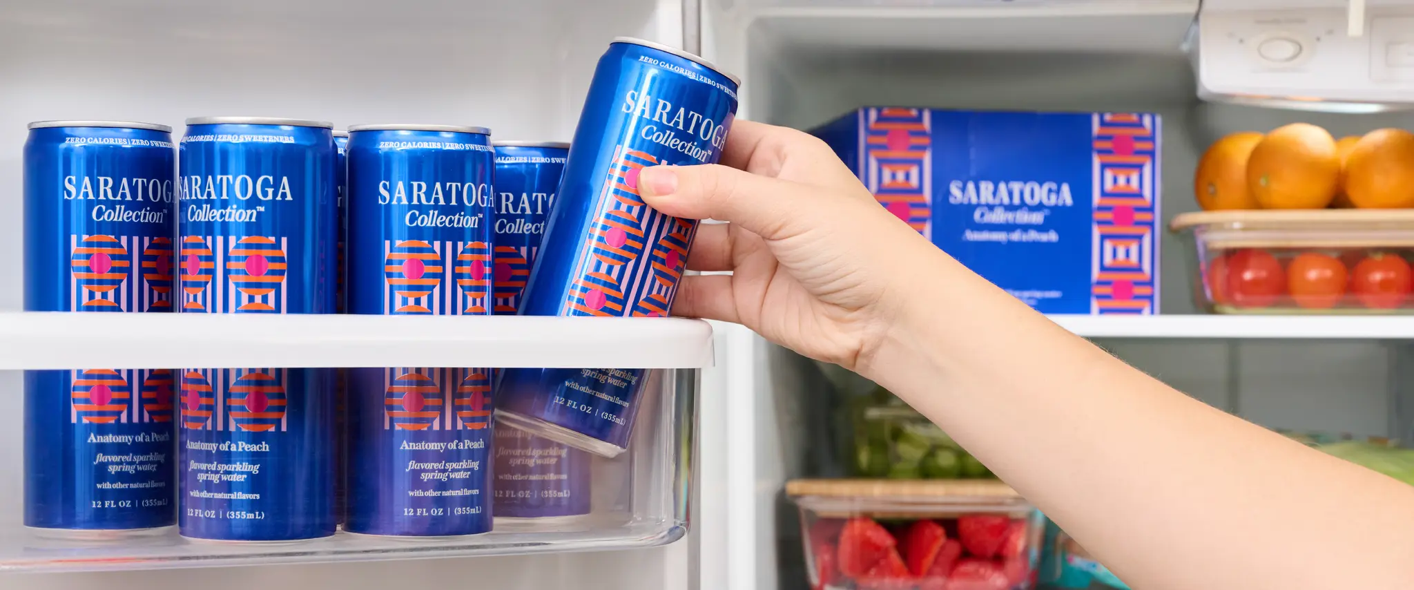 A hand reaching for a blue can of Saratoga sparkling flavored water from a refrigerator shelf, with more cans and assorted fruits visible inside.