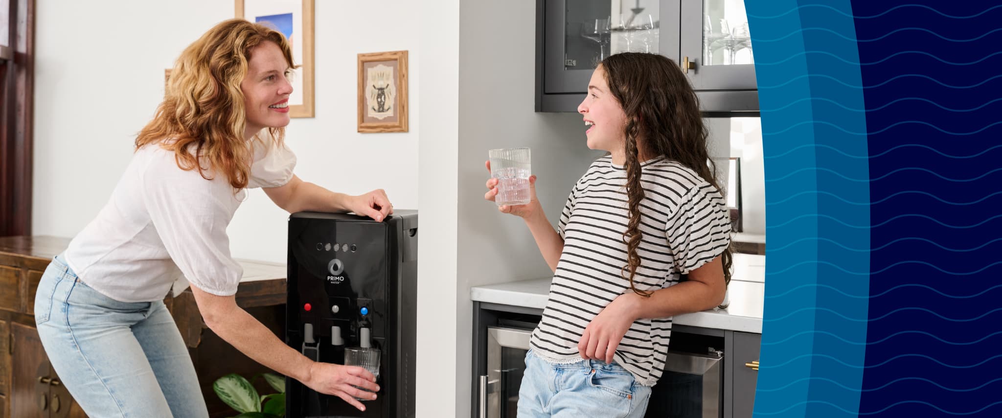 A woman fills a glass of water from a dispenser while a girl in a striped shirt and jeans leans against the counter, smiling with her own glass.