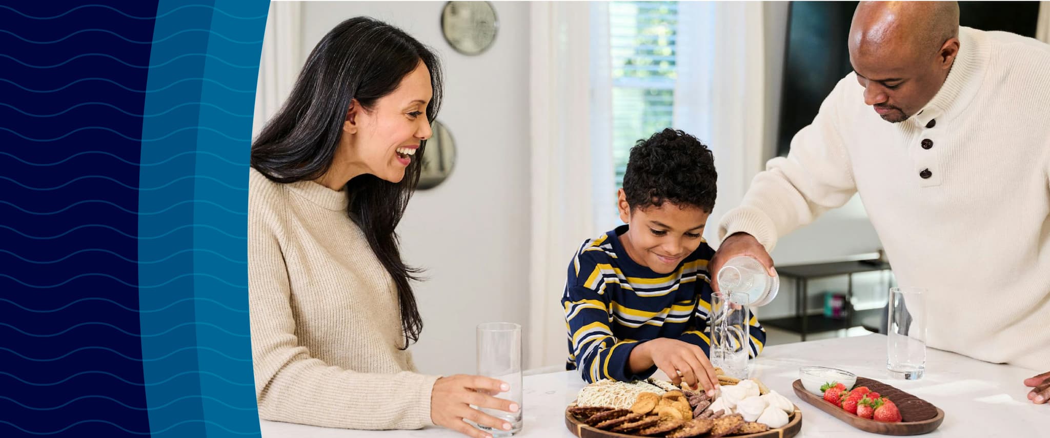 A family enjoys snacks at a kitchen counter; a man pours bottled water, while a woman and child smile, reaching for food on a wooden plate.