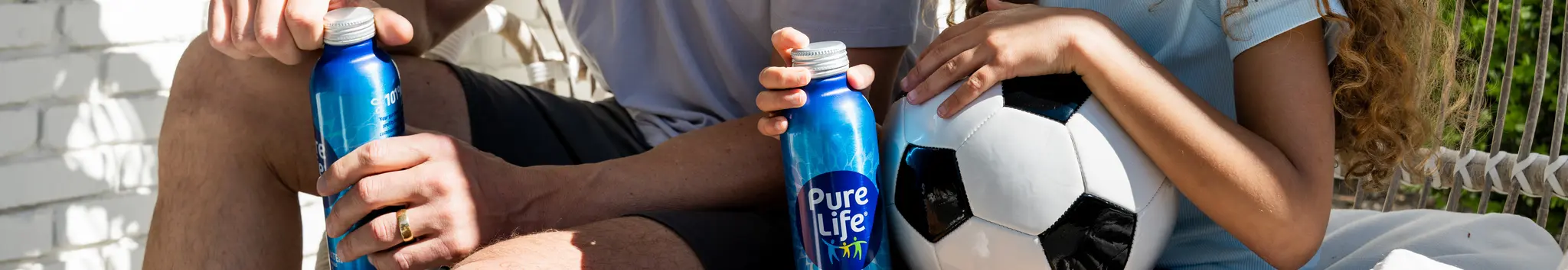 Two people sitting outdoors, holding blue Pure Life aluminum water bottles, with one person also holding a soccer ball.